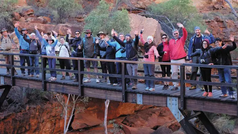 A happy group on a 3-Night Rock adventure smiles and waves from a wooden bridge in a scenic rocky canyon, enjoying the outdoors.