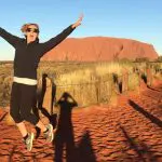 Excited woman leaps with arms raised on red dirt track in Macdonnell Ranges near Uluru, under vibrant blue sky, Australia adventure.