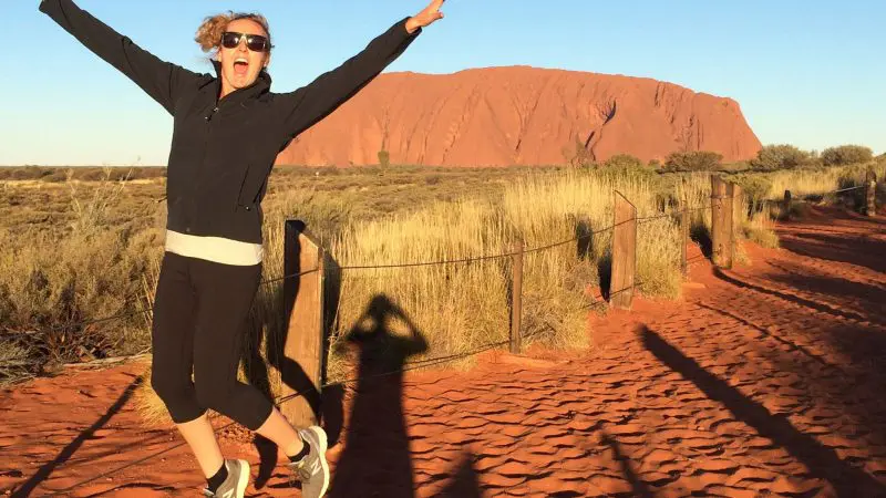 Excited woman leaps with arms raised on red dirt track in Macdonnell Ranges near Uluru, under vibrant blue sky, Australia adventure.
