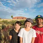 Three happy travellers smile together at Uluru, enjoying the iconic 3 Night Rock The Centre Yulara to Yulara adventure tour outdoors.