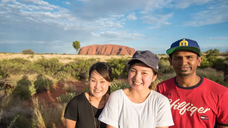 Three happy travellers smile together at Uluru, enjoying the iconic 3 Night Rock The Centre Yulara to Yulara adventure tour outdoors.