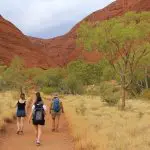 Three hikers trek the 3-Night Rock The Centre Yulara to Yulara trail amid striking red hills and scattered vibrant green trees.