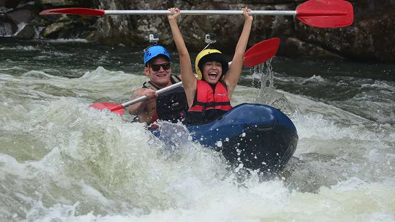 Adventurous duo white-water rafting near Mission Beach, one triumphantly lifts paddle whilst both smile, thrilling ride over choppy waves.