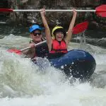 Adventurers in helmets and life jackets conquer rapids at Mission Beach, raising paddles high as water splashes during full-day rafting.