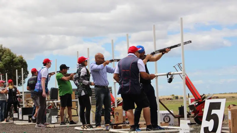 Participants at Have A Go Clay Target Shooting aim shotguns outdoors in red caps and waistcoats beneath an overcast sky, enjoying the event.
