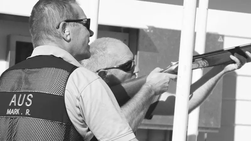 Two men in sunglasses, one wearing an AUS vest, aim a shotgun outdoors during a Have A Go Clay Target Shooting event, black and white.