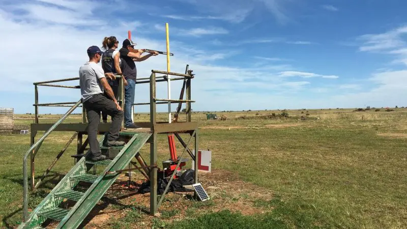 Three shooters on a raised platform at Vic Werribee aim at clay targets, enjoying a clear blue sky during competitive shooting.