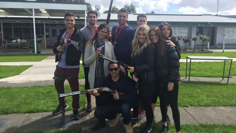 Eight happy participants with medals and shotguns pose after a Have A Go Clay Target Shooting event outdoors, celebrating their victory.