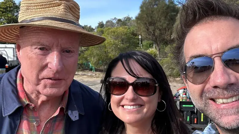 Three happy travellers enjoy a 1 Day Home Away Tour outdoors, wearing sunglasses and a straw hat, with trees and a clear blue sky.