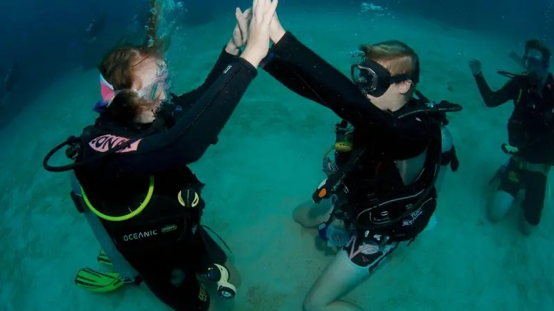Two scuba divers celebrate with an underwater high five during a 1–2 Day PADI Refresher Course as coursemates watch from the sea bed.
