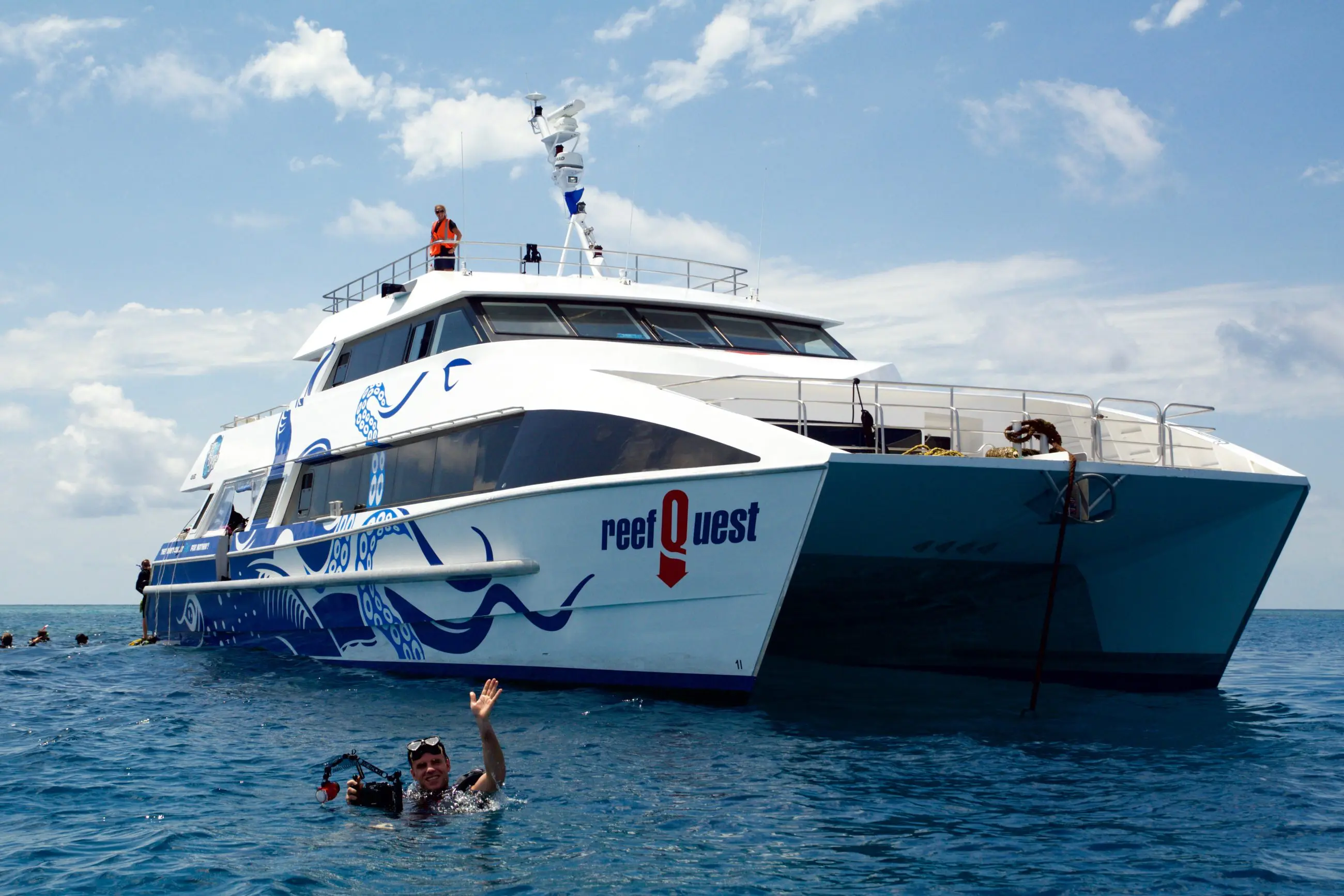 A man waves as he swims beside the Aquaquest Premium Day Trip boat on clear ocean waters beneath a scenic partly cloudy sky.
