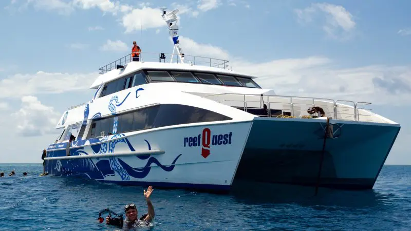 A man waves as he swims beside the Aquaquest Premium Day Trip boat on clear ocean waters beneath a scenic partly cloudy sky.