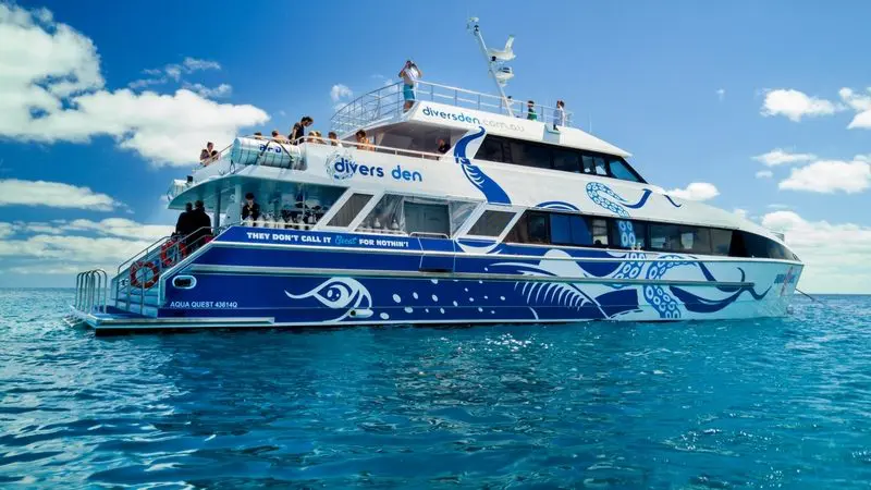The Aquaquest Outer Reef Snorkel Special catamaran glides on crystal-clear, tranquil water beneath a radiant blue, sunlit sky.