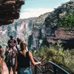 Walkers on a Blue Mountains day tour walk a scenic rocky path above a gorge, surrounded by lush views, sharing a picnic lunch.