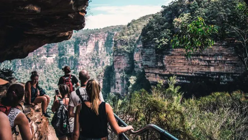 Walkers on a Blue Mountains day tour walk a scenic rocky path above a gorge, surrounded by lush views, sharing a picnic lunch.