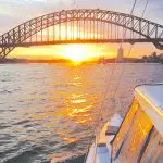 Experience a BYO Sunset Sparkle Catamaran Cruise as a luxury boat sails past the iconic Sydney Harbour Bridge at golden sundown.