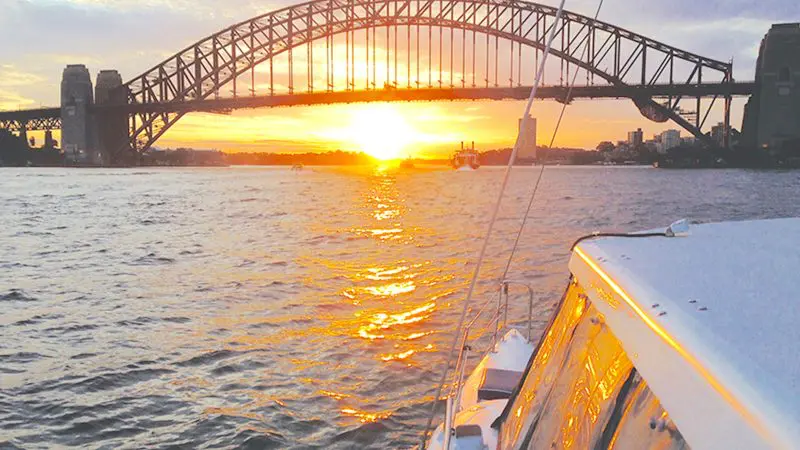 Experience a BYO Sunset Sparkle Catamaran Cruise as a luxury boat sails past the iconic Sydney Harbour Bridge at golden sundown.