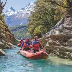 Adventurers in life jackets paddle a vibrant red raft down the crystal-clear Dart River, framed by rugged cliffs and majestic mountains.