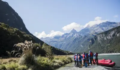 Adventurers in life jackets gather beside vibrant red Funyaks on the scenic Dart River, framed by majestic mountains and blue skies.