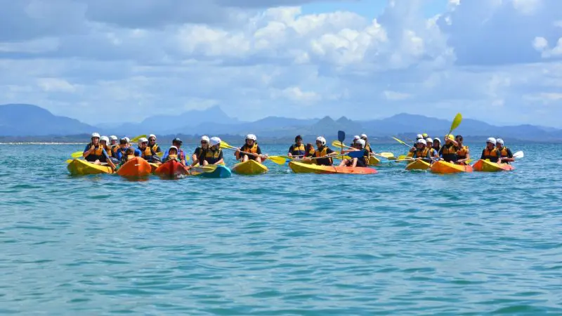 Adventurers in bright life jackets kayak on vibrant blue waters in Byron Bay, mountains scenic in the backdrop, on a guided tour.