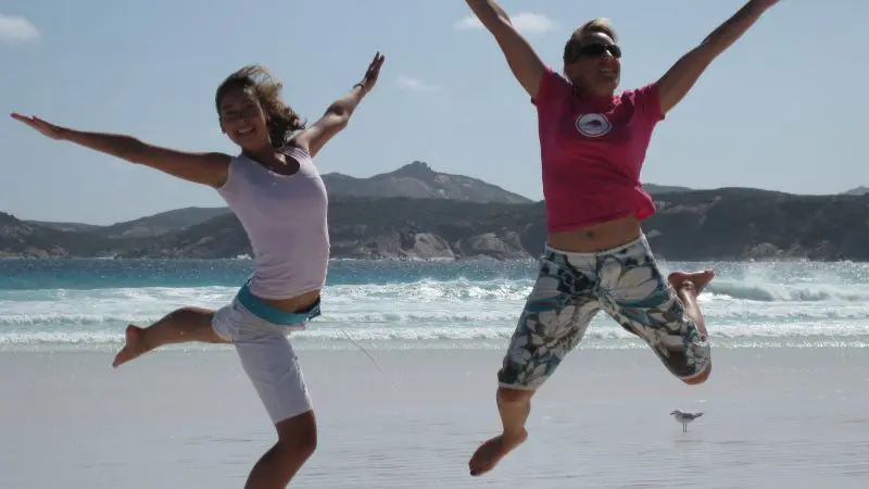 Two women leap with joy on a sunlit sandy beach during a 10-Day Broome to Perth West Coast Adventure, under clear blue skies.