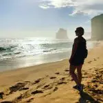 Woman stands on pristine sandy beach, admiring dramatic ocean cliffs in golden sunlight—Great Ocean Road Day Tour with Go West.