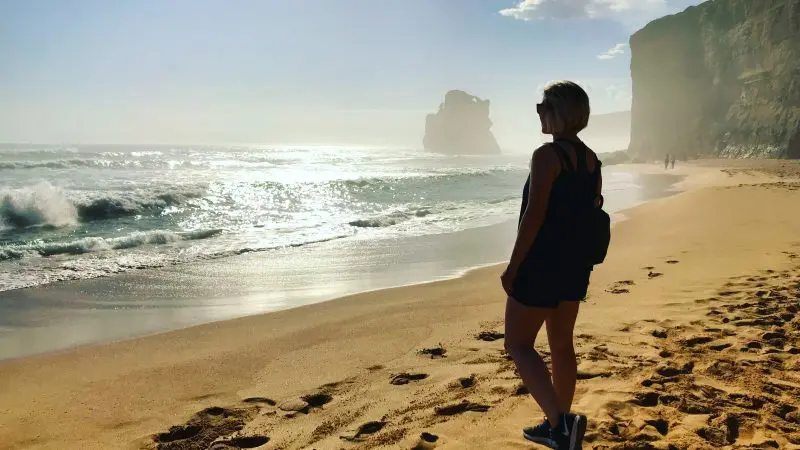 Woman stands on pristine sandy beach, admiring dramatic ocean cliffs in golden sunlight—Great Ocean Road Day Tour with Go West.