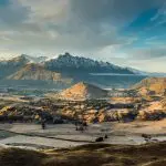 Breathtaking snow-capped peaks tower over a sunlit Queenstown valley, with lush fields and trees beneath a dramatic cloudy sky.