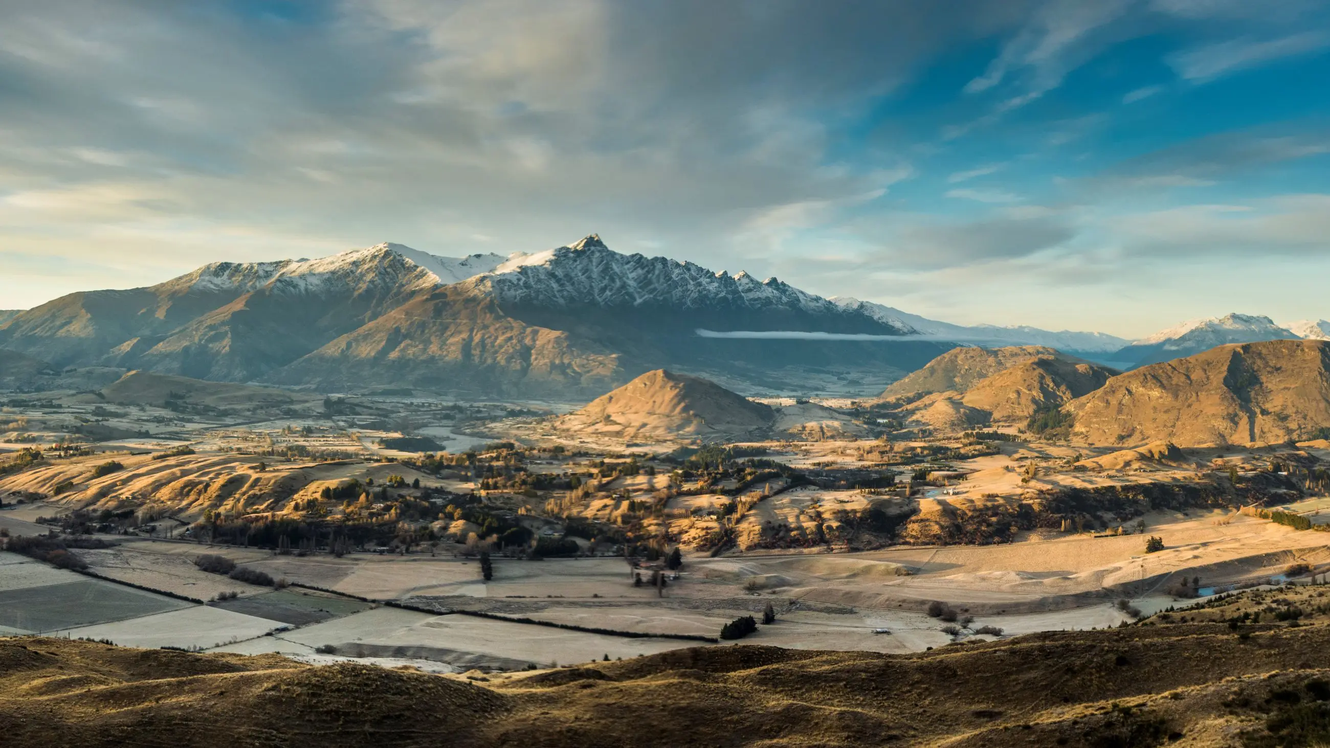 Breathtaking snow-capped peaks tower over a sunlit Queenstown valley, with lush fields and trees beneath a dramatic cloudy sky.