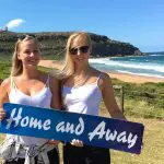 Two women in white tops display a Home and Away sign on Celebtime Tours beach, with cliffs and blue sky in the sunshine behind them.