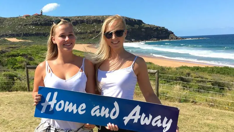 Two women wearing white tops hold a Home and Away sign on a sunny beach during a popular 1 Day Home and Away Location Tour.