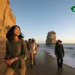 Four travellers stroll along a sandy beach at sunset by scenic cliffs on a Great Ocean Road 1 Day Sunset Tour; Bunyip Tours logo visible.
