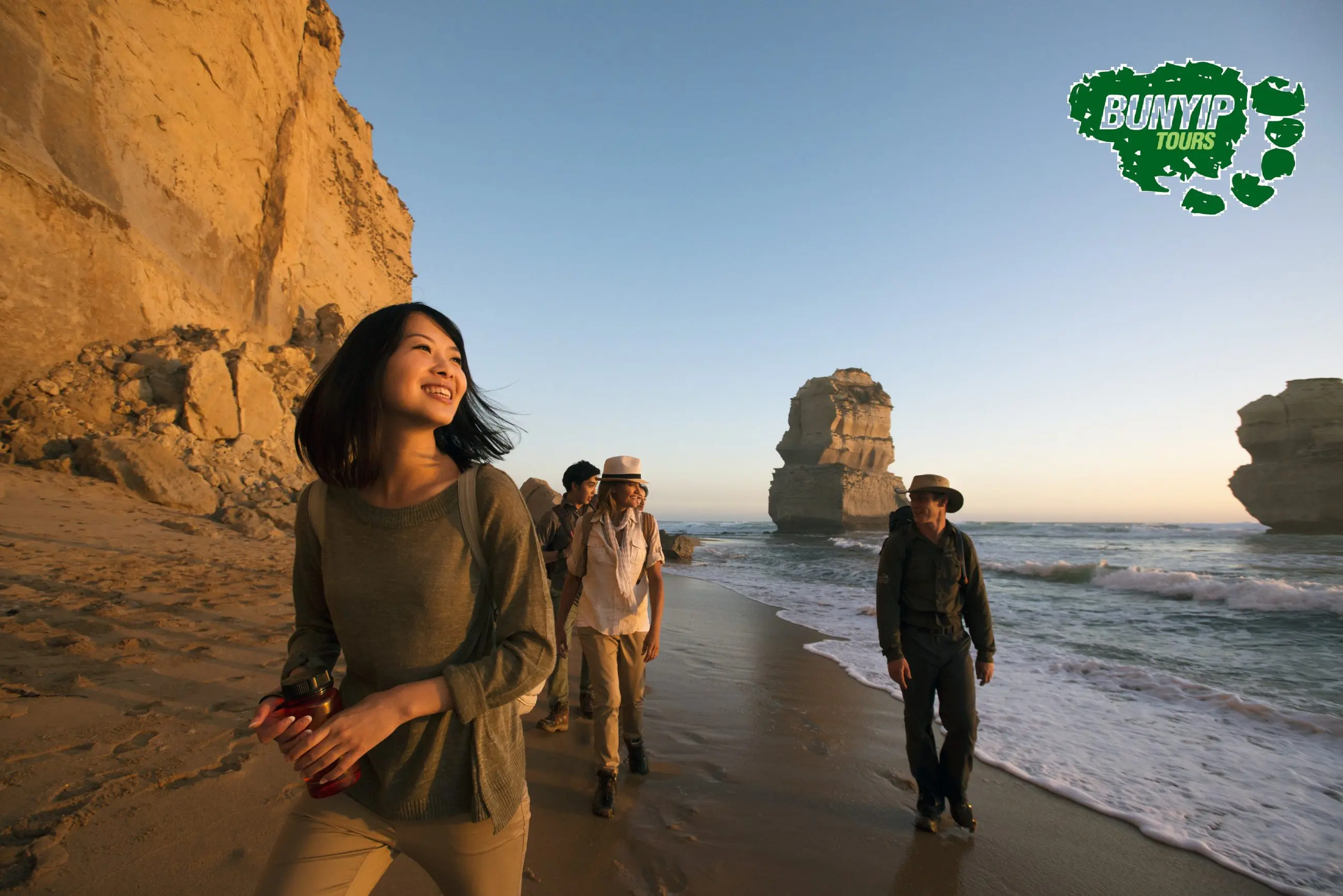 Four travellers stroll along a sandy beach at sunset by scenic cliffs on a Great Ocean Road 1 Day Sunset Tour; Bunyip Tours logo visible.