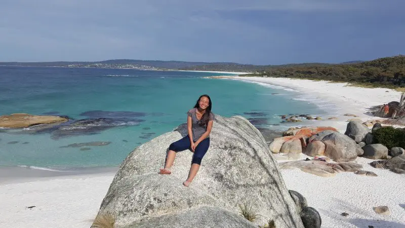 Happy woman beams atop a scenic rock beside crystal-clear turquoise waters on the 7 Day Super 7 Tasmania Tour with Under Down Under.