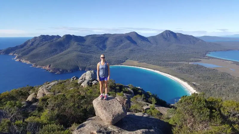 Woman stands atop a rocky outcrop, gazing over a stunning curved bay on the 7 Day Super 7 Tasmania Tour by Under Down Under Tours.
