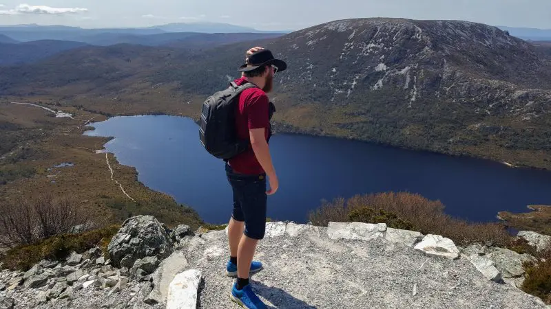 Traveller with a rucksack stands atop a rugged Tasmanian cliff, taking in breathtaking panoramic views from the 7 Day Super 7 Tour.