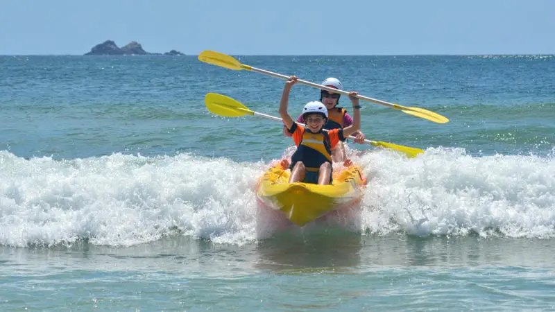 Two adventurers in life jackets and helmets paddle a kayak through sparkling ocean waves on a sunny Byron Bay Sea Kayak Tour.
