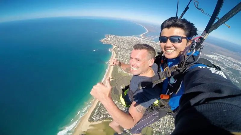 Experience the thrill of tandem skydiving over Sunshine Coast Beach—two men smile, give thumbs up, and soar above stunning ocean views.