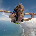 Thrilling tandem skydive above Sunshine Coast Beach, two people smiling and waving mid-air with scenic ocean views and clouds behind.