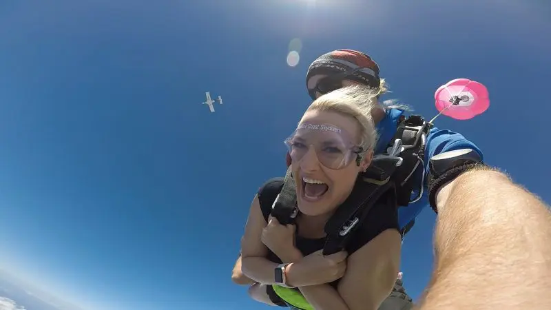 Woman smiling with excitement during Sunshine Coast Beach Tandem Skydive, aeroplane overhead, parachute open against clear blue sky.
