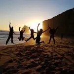 Energetic group silhouettes jumping on a sandy beach at sunset during Go West Tours’ 1 Day Great Ocean Road Sunset Tour, Australia.