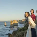 Three people enjoy a scenic sunset on a boardwalk during a 1 Day Great Ocean Road Sunset Tour with Go West Tours, breathtaking views.