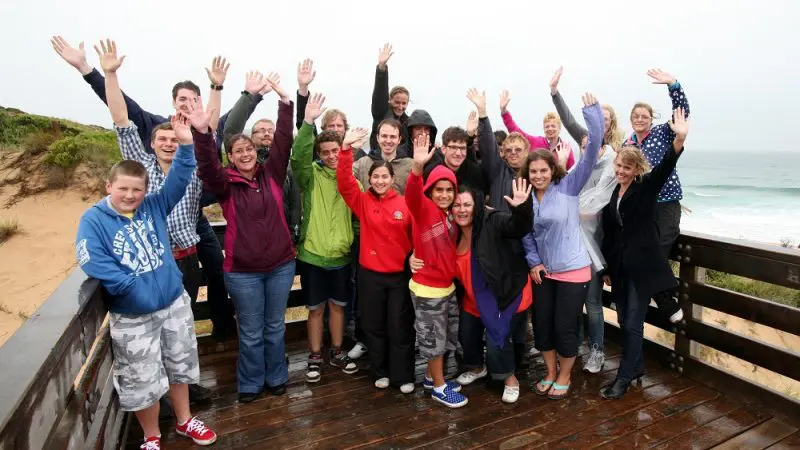 Tour group on wooden decking by Phillip Island beach smiles and waves during Penguin Parade Tour, enjoying scenic sea views.