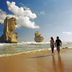 Couple walking by the iconic 12 Apostles limestone stacks on a Great Ocean Road day tour with Go West Tours beneath clear blue skies.