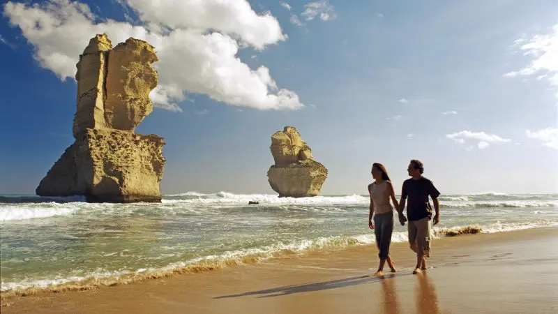 Couple walking by the iconic 12 Apostles limestone stacks on a Great Ocean Road day tour with Go West Tours beneath clear blue skies.