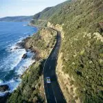 Experience the iconic 1 Day Great Ocean Road 12 Apostles Tour with Go West Tours as a car winds between lush green hills and the stunning sea.