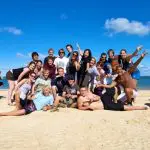 Group of happy travellers posing on a sunny East Coast Explorer tour, sandy beach, parked vehicles, and vibrant blue sky in background.