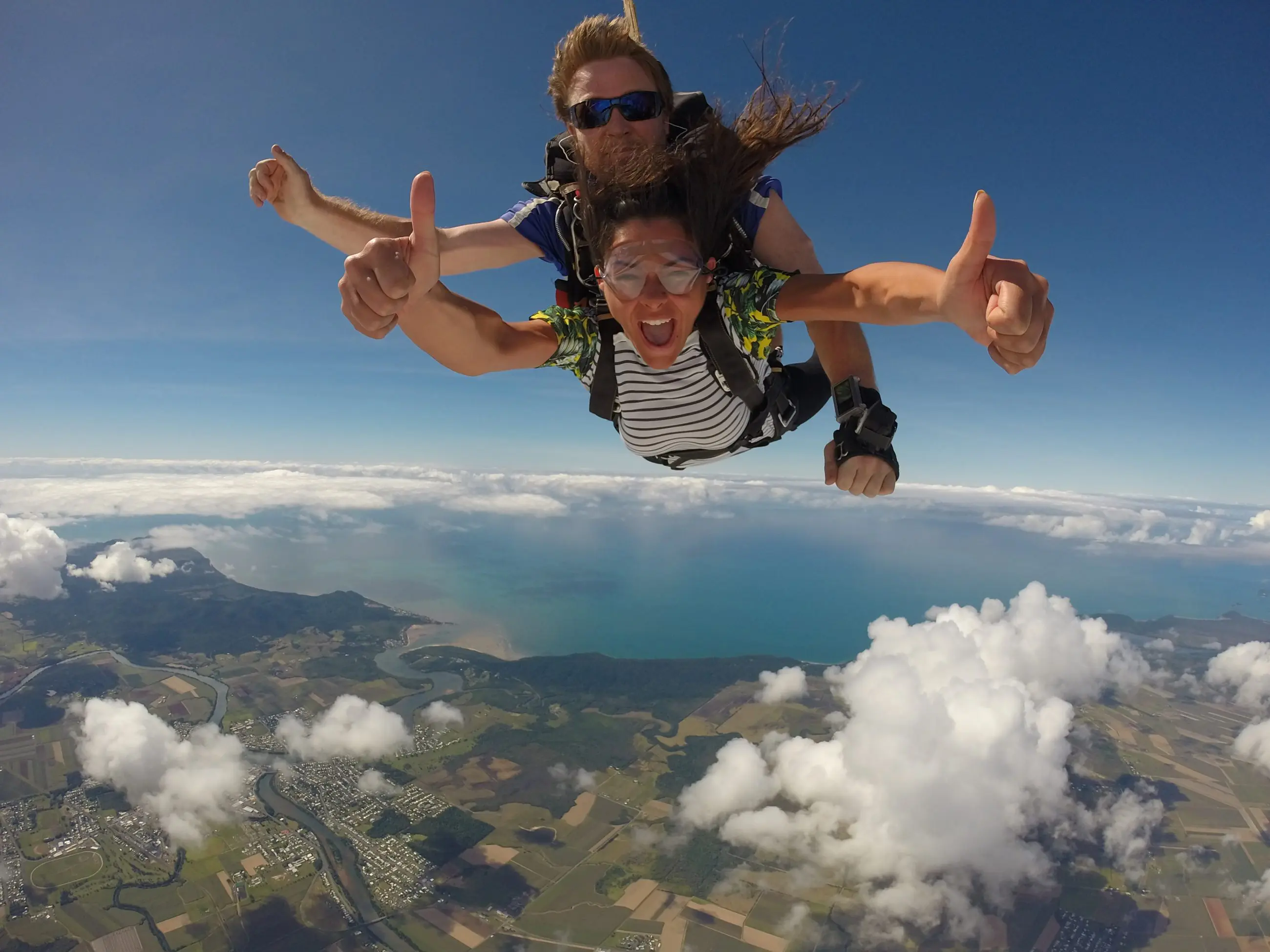 Two people tandem skydiving with Skydive Cairns, giving thumbs up high above tropical clouds and ocean, Great Barrier Reef below.