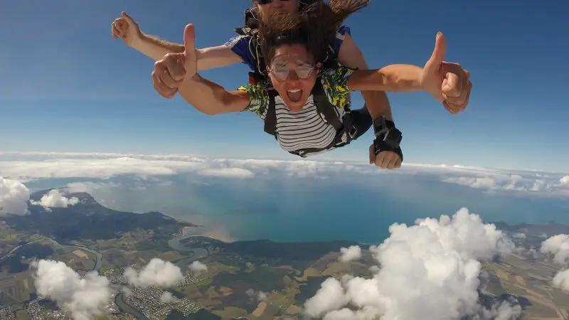 Two people tandem skydiving with Skydive Cairns, giving thumbs up high above tropical clouds and ocean, Great Barrier Reef below.