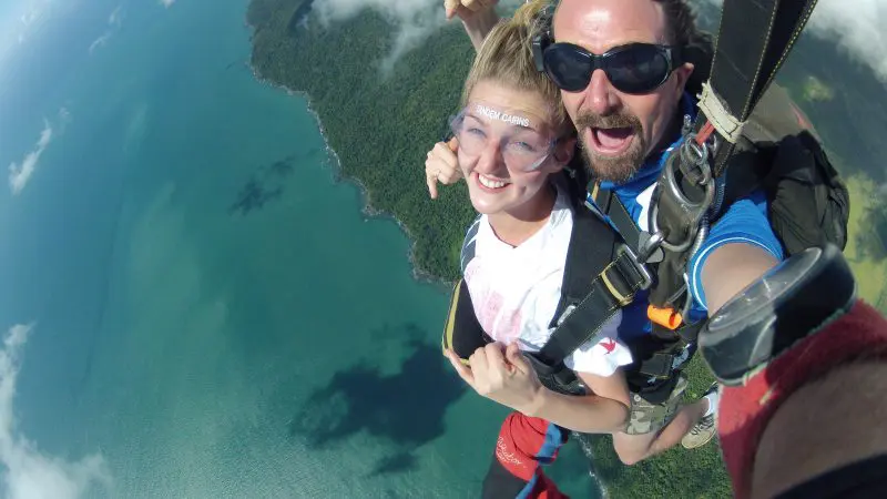 Tandem skydivers free-fall above scenic coastline, grinning at camera, with blue ocean waves and lush green landscape below.